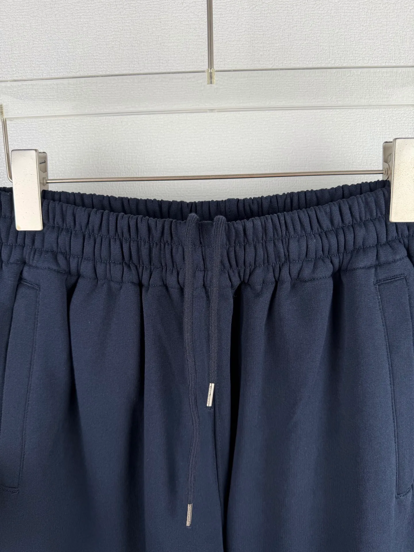 ACNE STUDIOS embroidered logo fleece sweatpants wide-leg trousers. Fabric: 32-count double yarn top + 16-count terry bottom yarn G The veil is made of 32-count fine yarn. The high-count yarn is smooth and delicate. After weaving, it forms a smooth and compact surface. It not only resists pilling and wear-resistant, but also gives the fabric a touch as delicate as skin. The base yarn is made of 16-count roving. The low-count yarn is fluffy and hairy. It naturally forms a rich terry layer during the weaving process, providing a sufficient source of fiber for subsequent sanding. This combination of fine surfaces and thick soles strikes a perfect balance between warmth and skin-friendliness. Mechanical sanding is the soul process of this fabric. By rubbing the bottom surface of the roller wrapped with emery leather at high speed, the fiber ends of the 16-count roving are evenly pulled out and cut off, forming a dense and uniform short pile layer. The smoothness and neatness of needle embroidery and the soft velvet feel of brushed fabrics form a material dialogue between hardness and softness. The embroidery area is smooth and crisp, with sharp lines, like delicate inscriptions carved on a rich suede canvas; while the surrounding brushed fabric presents a soft matte natural texture. The spacing between stitches is consistent and the strength is balanced to ensure that each stitch is placed accurately. This rigorous stitching structure makes the edges of the logo present a sharp and clear outline, and the strokes of the text are full and smooth. Even the curved turning points of the floral fonts can achieve a smooth transition through the connection of tight stitches.